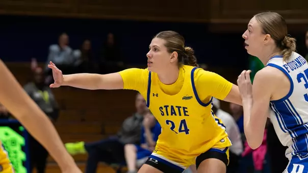 A women's basketball player in a yellow uniform defends against a player in a white uniform