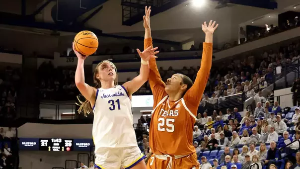 A woman in a white basketball uniform goes up for a shot while a player in an orange uniform defends