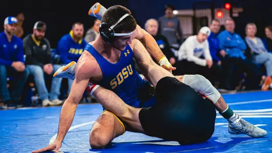 A man in a blue singlet attempts to takedown another man wearing a black singlet from behind