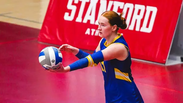 A women in a blue volleyball jersey prepares to serve a volleyball during a match. A red volleyball court and red Stanford sign are visible in the background.