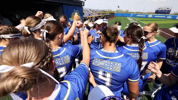 Members of the team huddle in front of their dugout with their hands up in the air