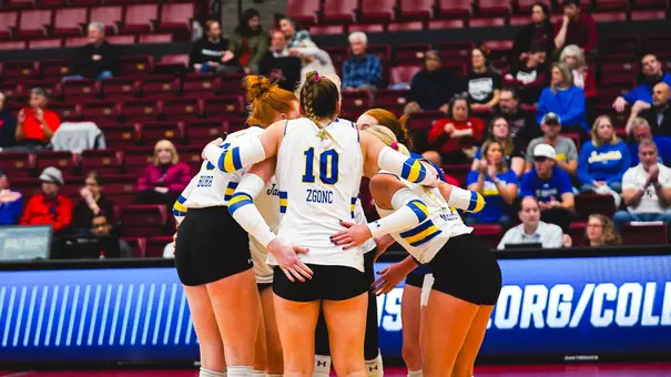 A women's volleyball team huddles on the court with red seats in the background