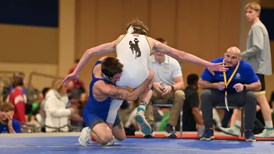 A man, Alek Martin, takes down another man wearing a white singlet in a wrestling match.