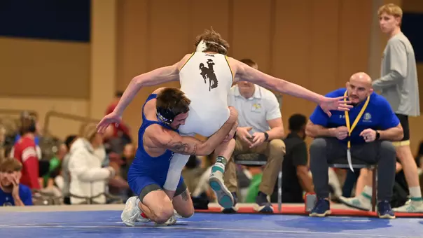 A man, Alek Martin, takes down another man wearing a white singlet in a wrestling match.