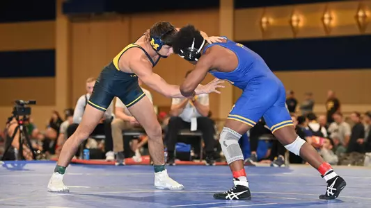 A man in a blue singlet interlocks with another man, in a dark blue singlet, during a wrestling match.