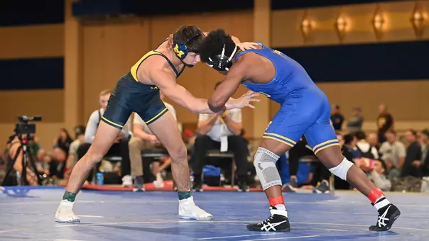 A man in a blue singlet interlocks with another man, in a dark blue singlet, during a wrestling match.