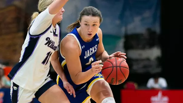 A women's basketball player in a blue uniform dribbles a basketball while a player in a white uniform defends