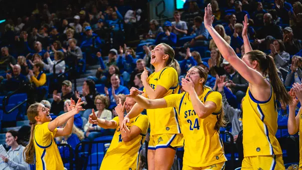 SDSU women’s basketball players in yellow uniforms on the bench cheer and raise their arms during a play as the home crowd claps behind them.