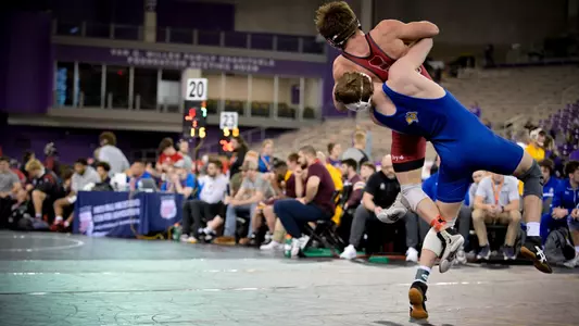 A wrestler in a blue singlet, Cael Swensen, picks up his opponent a wrestler in a red singlet during a match