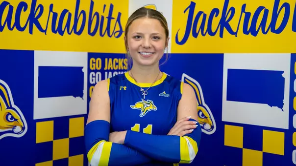 A women's volleyball player in a blue uniform, Anna Johannson, poses with crossed arms in front of a branded South Dakota State backdrop.