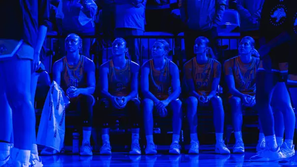 Basketball team seated on the bench during starting lineup announcements with coaches and players standing nearby, with a crowd of fans watching from the stands under blue arena lighting.