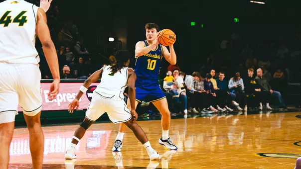 A men's basketball player, Kalen Garry, holds a basketball with a defender in a white uniform guarding him
