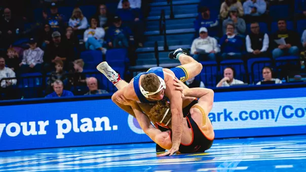 A wrestler in a blue singlet grabs the legs of a wrestler in a black singlet during a wrestling match.