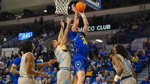 A basketball player in a blue uniform, Alec Squires, jumps toward the hoop for a shot while several defenders in gray uniforms attempt to block him during a game in a crowded arena