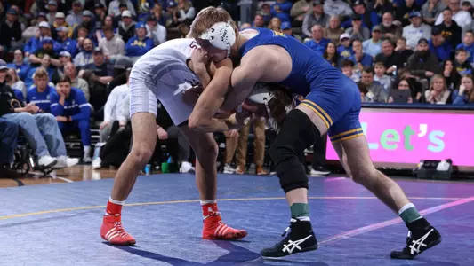 A wrestler in a blue singlet, Cale Seaton, holds a headlock against a wrestler in a white singlet during a wrestling match