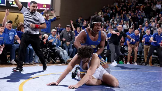 A wrestler in a blue singlet looks up while on top of another wrestler in a white singlet in a wrestling match