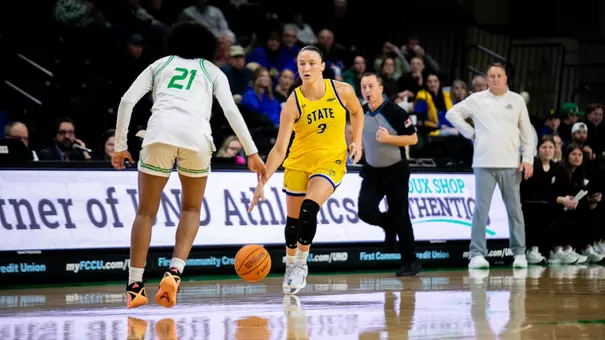 A women's basketball player in a yellow uniform, Maddie Mathiowetz, dribbles a basketball up the court toward a defending player in a white uniform.