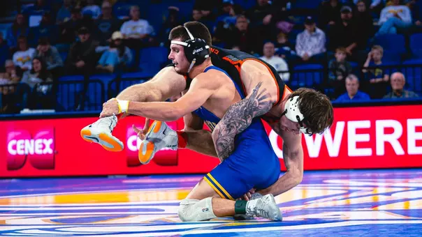 A wrestler in a blue singlet picks up a wrestler on his shoulders during a match.