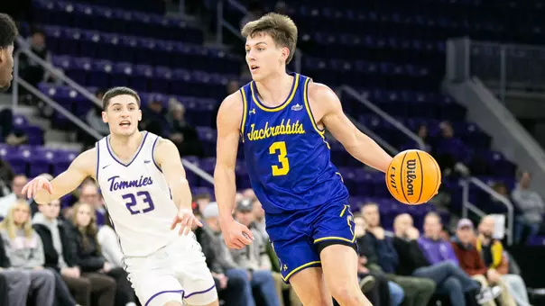 A men's basketball player in a blue uniform, Joe Sayler, dribbles a basketball with a defender in a white uniform following him