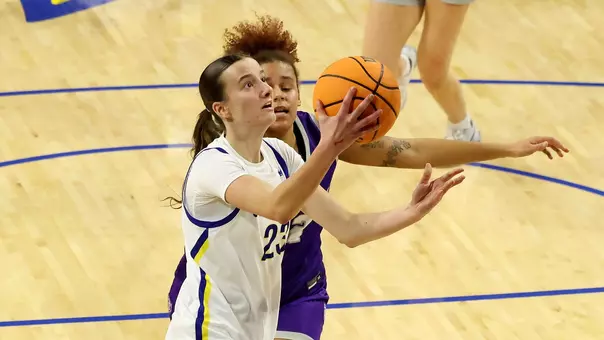 A women's basketball player in a white uniform, Katie Vasecka, goes up for a layup during a game. A defensive player in a purple uniform is visible behind her.