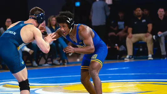 A wrestler in a blue singlet, grapples with another wrestler in a navy singlet during a wrestling match.