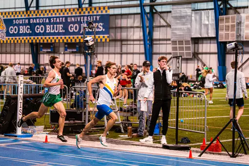 Men's runner in white and blue races around the curve of the track with a competitor in white and green on his heels