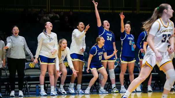 SDSU women’s basketball players on the bench stand and cheer as a teammate makes a 3-pointer on the court.