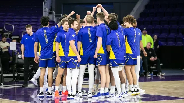 A group of South Dakota State basketball players in blue and yellow uniforms stand in a tight huddle on the court with arms raised
