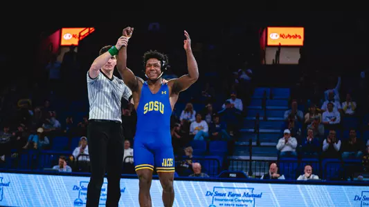 A wrestler in a blue singlet has hand raised by a referee after a win.