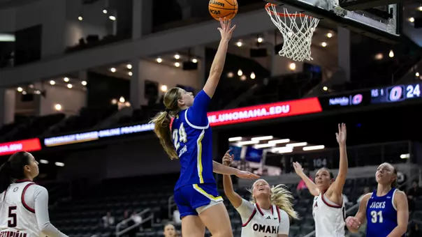 A women's basketball player in a blue uniform goes up for a layup while players in white uniforms look on.