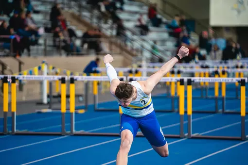 A male track athlete dips his head with his arms back, finishing the 60-meter hurdle race