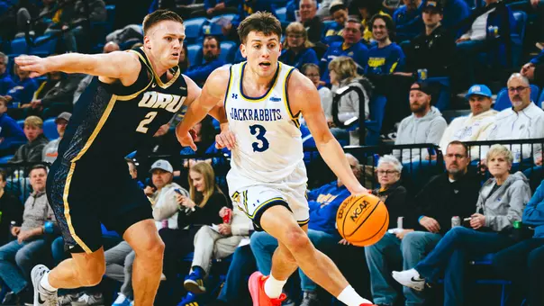 A men's basketball player, Joe Sayler, in a white uniform dribbles a basketball with a defender alongside him