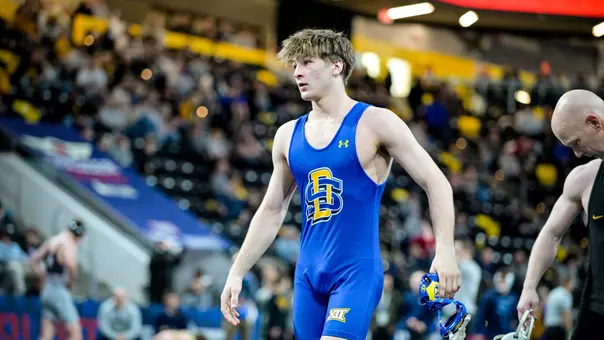 A wrestler in a blue singlet walks off the mat.