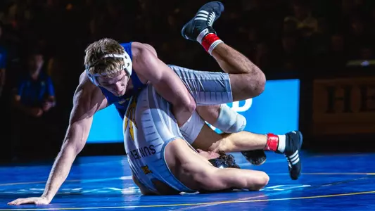 A wrestler, Cael Swensen, puts a wrestler in a white singlet's shoulders to the mat during an amateur wrestling match
