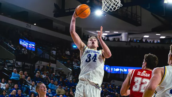 Basketball player, Damon Wilkinson, in a white Jackrabbits jersey attempts a shot near the hoop while a defender in a red jersey looks on during a game