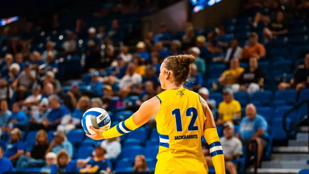 A volleyball player in a yellow uniform prepares to serve during a mathc.
