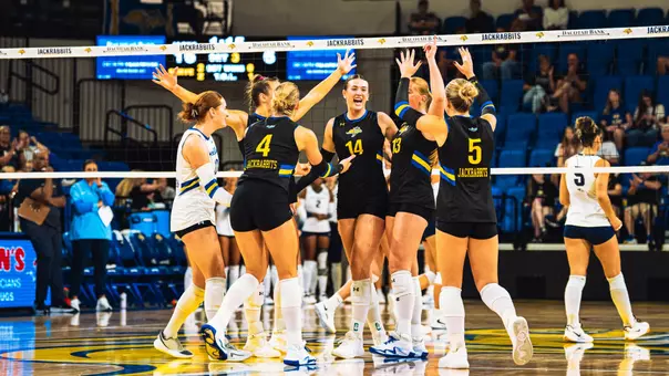 A group of volleyball players in black uniforms gathers around Hannah Mlsna wearing #14.
