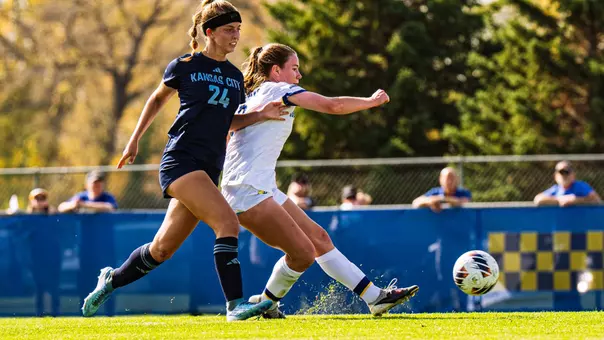 A woman in a white soccer uniforms kick a soccer ball with her left foot for a goal. An opposing player in a dark uniform closely follows behind.