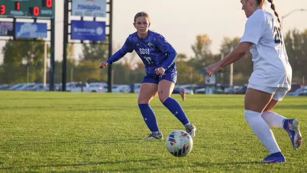 A woman in a blue soccer uniform looks on at a player with the ball at her feet as she defends her from a distance