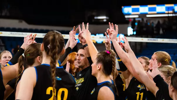 A group of volleyball players and coaches puts up bunny ears in a huddle after a win.
