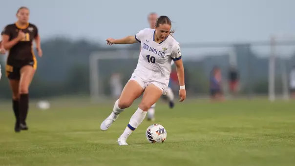 A woman in a white soccer uniform looks down at a soccer ball while she prepares to run on a field. Players stand looking at her in the distance.