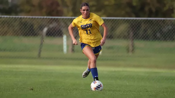 A woman in a yellow jersey with blue shorts chases after a soccer ball while running