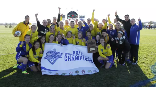 A team of women's soccer players pose with a championship trophy with a scoreboard behind them in the distance