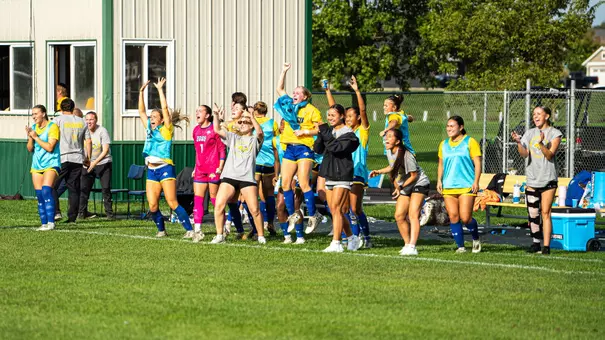 A group of women celebrate a play on a soccer field while standing in front of a press box along a bench area