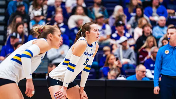 A volleyball player in a white uniforms stands with her hands on her knees.