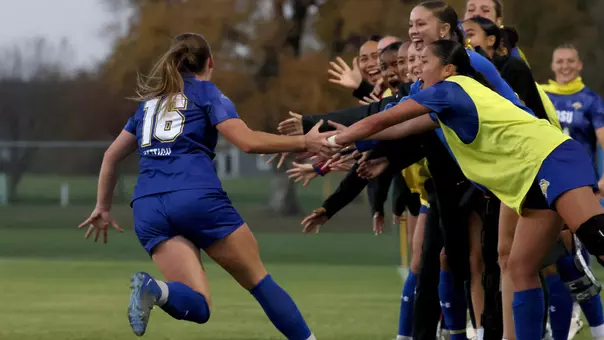 A woman in a blue soccer uniform runs up to teammates on the sideline and gives them fives along their sides