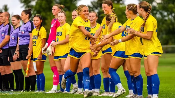 A photo of a soccer team in yellow and blue uniforms during starting lineup on the middle of the field. One player runs through a line of teammates slapping hands and smiling.
