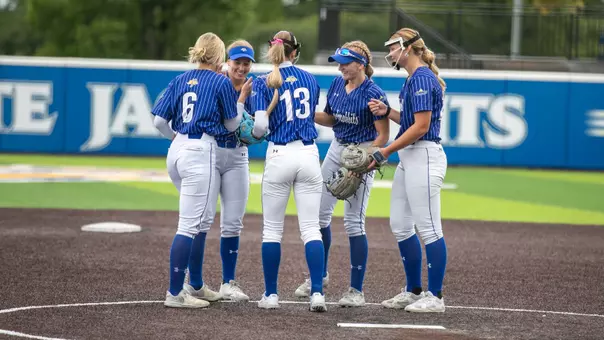 A group of five softball players in blue and white uniforms slap hands while smiling in the pitcher's circle of a softball diamond