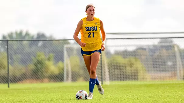 A woman in a soccer uniform, featuring a yellow jersey and blue shorts, looks down the field towards teammates with a soccer field at her feet