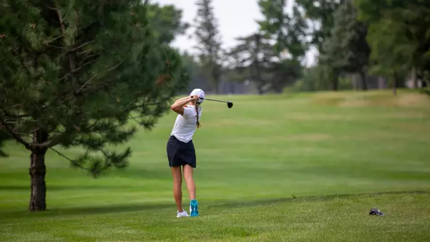 A golfer in a white cap and polo hits a ball in the fairway.